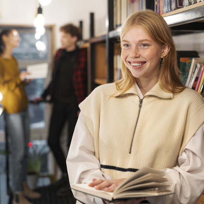 Une étudiante souriante tenant un livre dans une bibliothèque moderne, avec d'autres étudiants flous en arrière-plan, illustrant l'excellence académique et l'expérience étudiante chez HEC Paris.